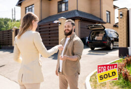 Female realtor in suit passing house key to bearded man and congratulating him with buying cottage house